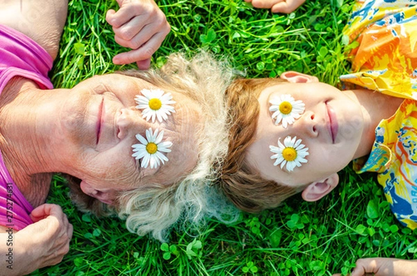 Obraz Happy family - grandmother with grandson having fun lying on the lawn with daisies on their eyes. Summer vacation with my grandmother. Family relationships and friendship