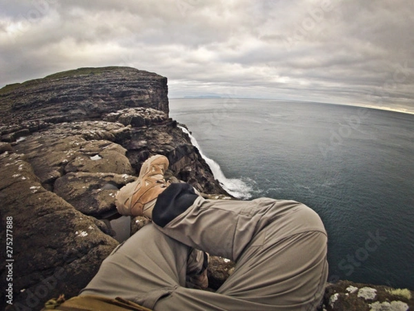 Fototapeta Tourist man sitting on a rock and looking at the endless sea. Wearing grey sport touristic pants, tan\beige army trekking boots.  Scenic waterfall in front of him, grey cliff and blue ocean on sides.