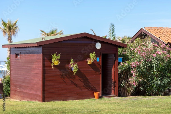Fototapeta Little brown painted shed in the middle of a garden with green grasses and trees
