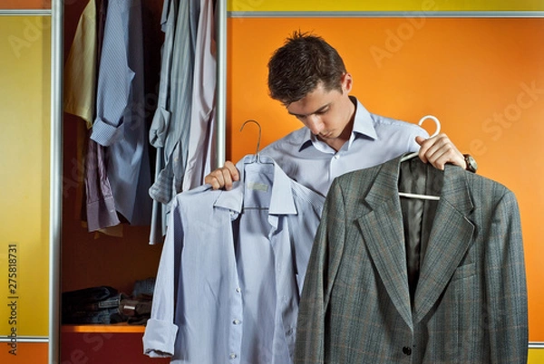 Fototapeta A man in a striped shirt tries clothes on the background of the closet. Yellow and orange wardrobe. The guy picks up the suit. The concept of clothing selection problem.