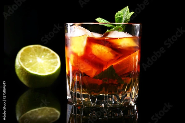 Obraz Glass of Dark Rum Cocktail with lime, orange, ice cubes and mint leaves on black mirror background.