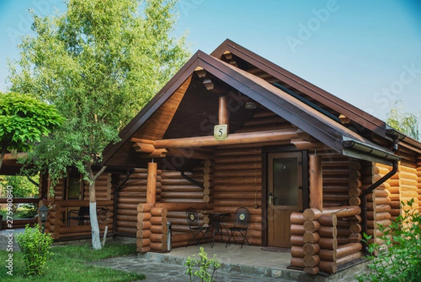 Fototapeta Wooden cottages with log and paved with wild stone paths on the background of green trees and flowering shrubs