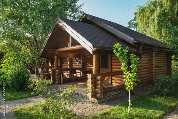 Fototapeta Wooden cottages with log and paved with wild stone paths on the background of green trees and flowering shrubs