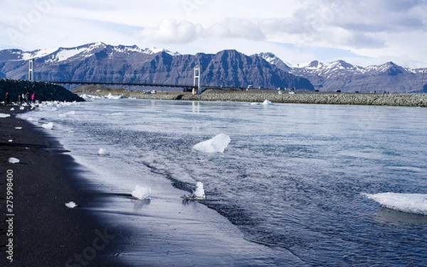 Fototapeta Iceland summer - Jokulsarlon Diamond Beach. Icebergs flowing into Atlantic, some washed up on the black sand beach. Picture taken from the sea towards the lagoon and inland mountains. Bridge and cars 