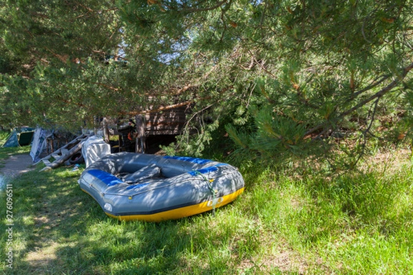 Fototapeta Tent camping in the shade under the trees with a wooden house and a large inflatable rafting boat near the river in the Altai Mountains. Extreme sports and recreation.