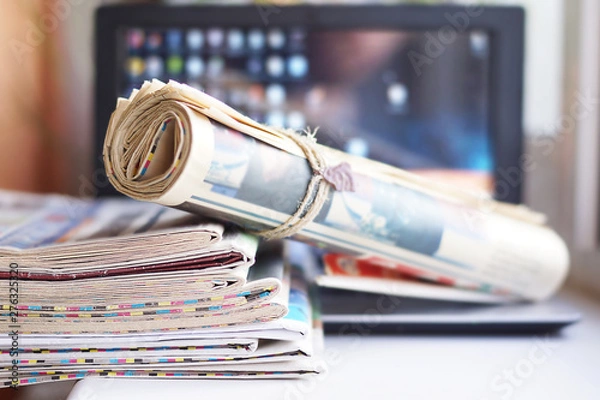 Fototapeta Stack of newspapers and laptop. Daily journals with news and personal computer. Tabloid papers with headlines  and articles and desktop on screen of electronic device. Different sources of information