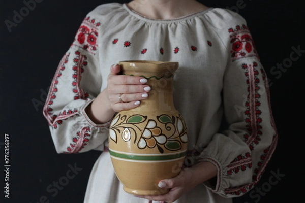 Obraz Closeup of part of a woman in Ukrainian embroidery with an earthenware jug in her hands on a black background