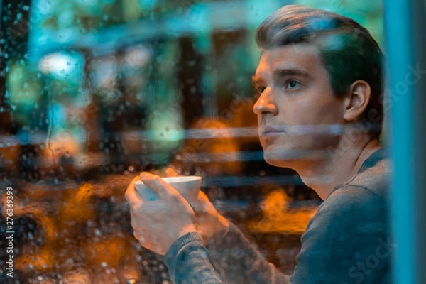 Obraz Handsome man drinking tea or coffee on rainy day and looking through the window