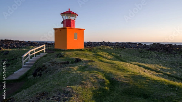 Fototapeta Summer Iceland - lighthouse on west cost near midnight. Low sun illuminating a red lighthouse close to rocky Atlantic shore. Green vegetation, blue sky above