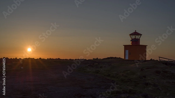 Obraz Icelandic  sunset - view of low sun above Atlantic with a lighthouse in foreground. Back light (close to midnight) throws beams on a rocky coastline dominated by a red land orange lighthouse.