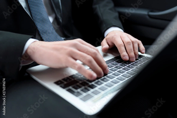 Obraz View of hands of businessman in a suit and tie, who is sitting in a car, with laptop on knees and typing on keyboard.