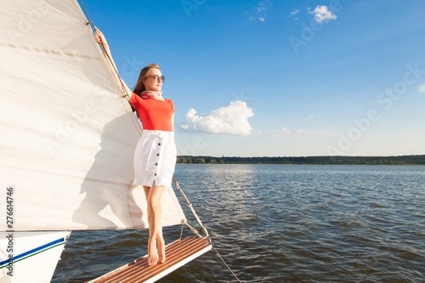 Fototapeta Model in a red blouse, white skirt, glasses, a red scarf on his head, standing on a white yacht, overlooking the sea and the blue sky, with a place for the inscription. The concept of luxury.