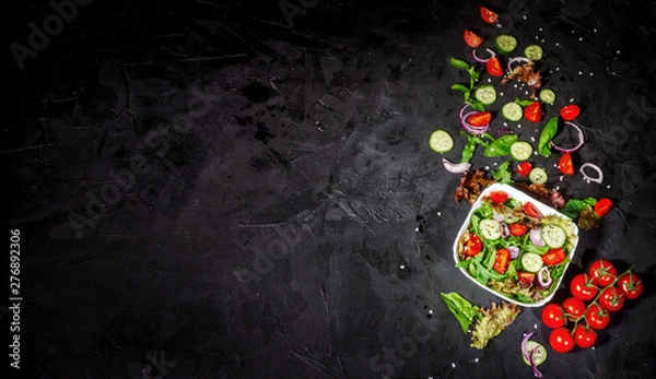 Fototapeta Various fresh mix salad leaves with tomato and cucumber in a glass bowl on dark background with ingredients. Top view with copy space
