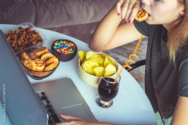 Fototapeta Girl works at a computer and eats fast food. Unhealthy food: chips, crackers, candy, waffles, cola. Junk food, concept.