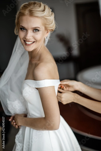 Fototapeta A young bride is looking at the camera while a bridesmaid is tying her corset.