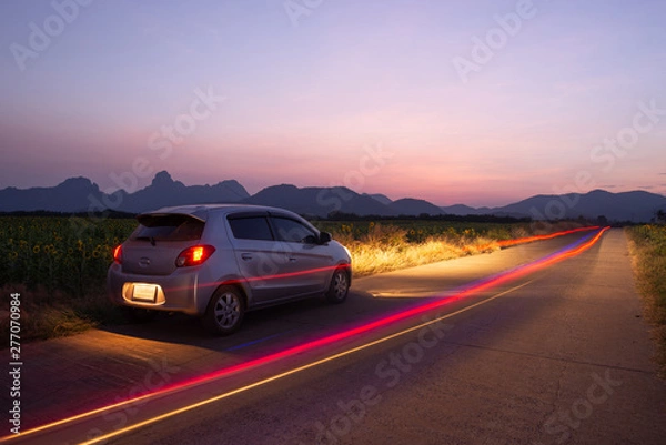 Fototapeta Travel car is parking at the road country side with landscape view beautiful sunset and light trails.