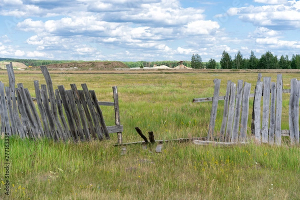Fototapeta old wooden rickety fence. Abandoned field with an old wooden fence. Old broken wooden fence. Summer sunny day with blue sky and white clouds. the effects of the hurricane. Disaster