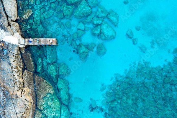 Obraz An aerial view of the beautiful Mediterranean Sea, with a wooden pier and a rocky shore, where you can see the textured underwater corals and the clean turquoise water of Protaras, Cyprus