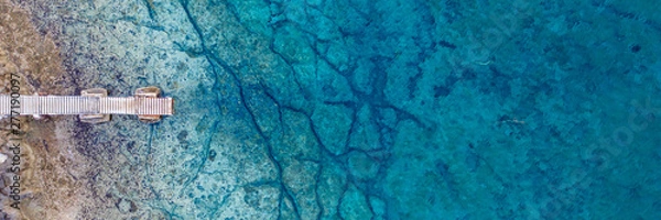 Obraz An aerial view of the beautiful Mediterranean Sea, with a wooden pier and a rocky shore, where you can see the textured underwater corals and the clean turquoise water of Protaras, Cyprus
