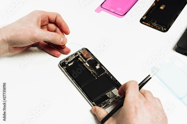 Fototapeta Smart Phone Battery Change Repair Maintenance. Repairman Hand Fix Smartphone Back Part Disassemble with Tweezers. Mobile Cellphone Assembly. Technician Work at White Workplace Copy Space View