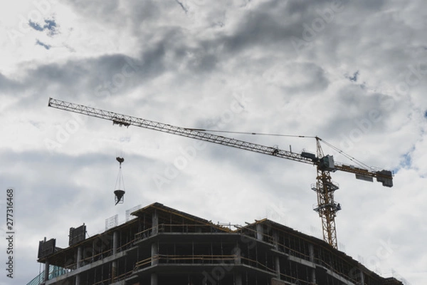 Fototapeta Building under construction against white cloudy sky. Big crane at the top at the time of work. Bottom view. Copy space.