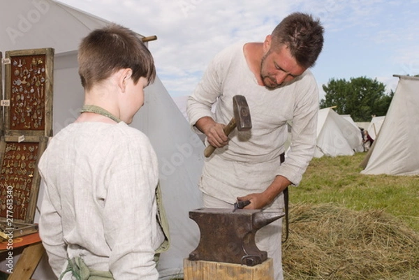 Fototapeta Training in metal forging. A blacksmith teaches a boy for a forge at a historic summer festival.