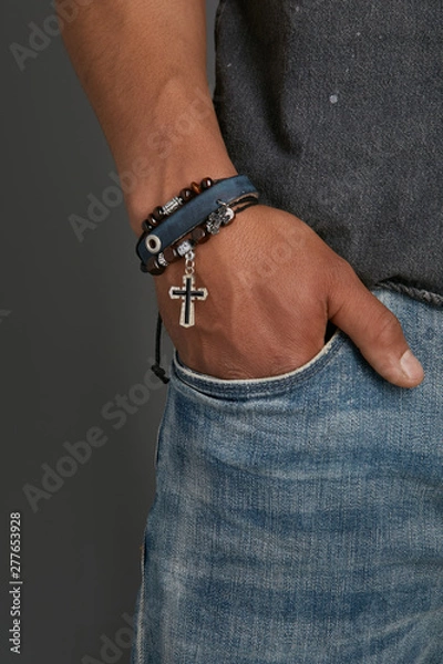 Fototapeta Cropped top and bottom photo of a man with a bracelet on his arm. The model on a gray background is dressed in a gray shirt and jeans. You can see a multi-layer bracelet made from leather and laces wi