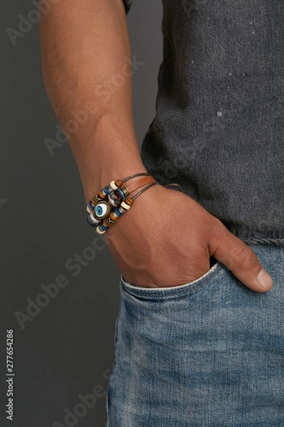 Obraz Cropped top and bottom photo of a man with a bracelet on his arm. The model on a gray background is dressed in a gray shirt and jeans. You can see a multi-layered bracelet made from leather and laces 