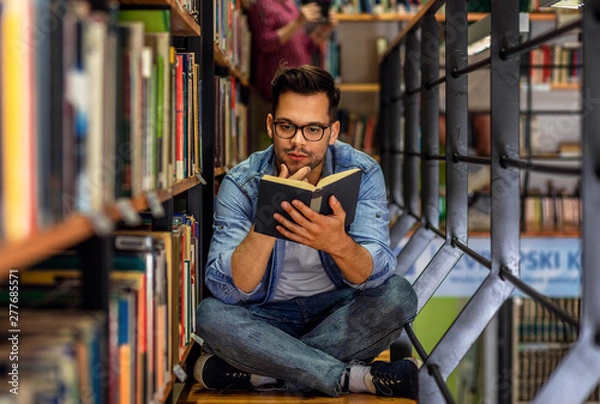 Fototapeta Young male student study in the library reading book while sitting near bookshelf.