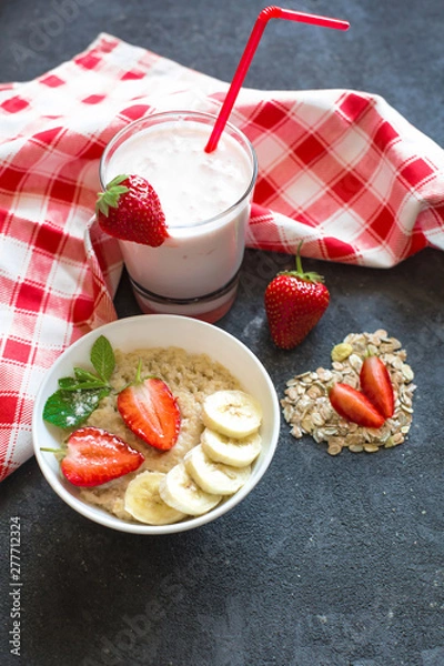 Fototapeta Porridge with strawberries and banana slices in a white plate, yogurt in a glass stand on a dark concrete background. Decoration of dry oatmeal and strawberry cloves.