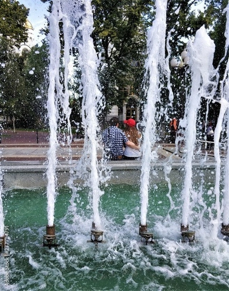 Obraz In the foreground is a photo of a fountain with spouting cascades of water, and through a spray of water you can see a couple in love where a guy hugs a girl.
