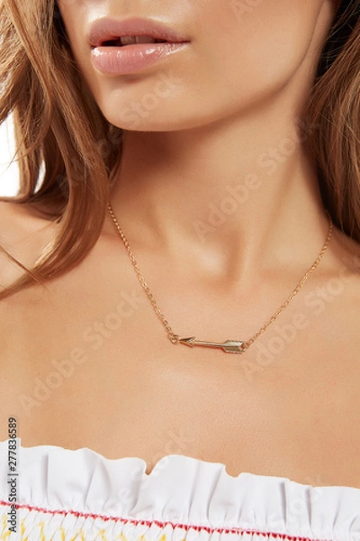Fototapeta Cropped shot of lady with plush lips, wearing golden necklace, decorated with pendant in view of an arrow. The girl with chestnut hair locks is turning head to side while posing on white background.