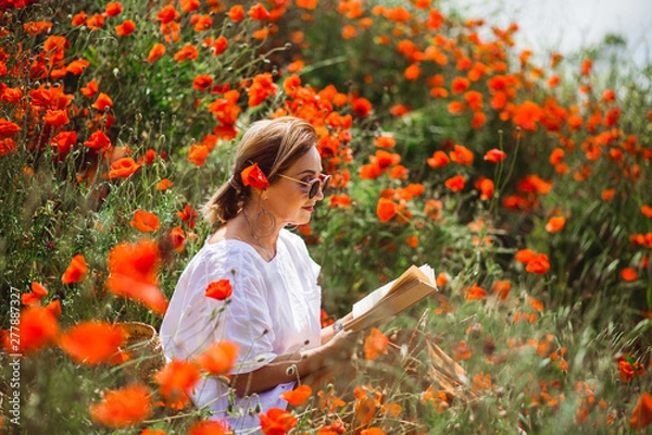 Fototapeta Woman reading a book in a field of red poppies in spring flowers mood.