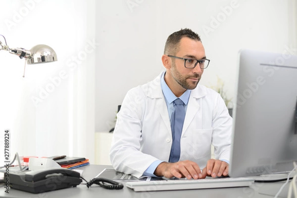 Fototapeta portrait of a handsome man male doctor in medical practice office working on computer