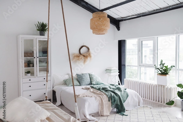 Fototapeta Modern scandinavian sunny bedroom with plants , floral pattern bedding and pillows. Space with white walls and wide window. Eco decor. Real photo.