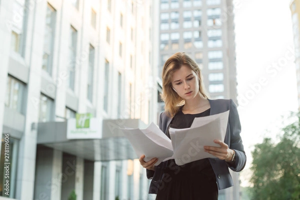 Obraz Concentrated caucasian financial adviser viewing business papers outdoors. Busy young businesswoman examining data in documents. Report concept. Office worker reading the documents on the street.