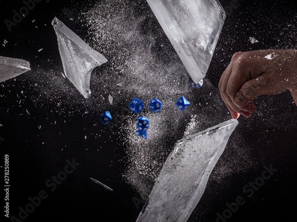 Fototapeta Blue Role Playing Dice Exploding through a Sheet of Ice - with a Caucasian Man's Hand to the Side Tossing the Dice and a Black Background