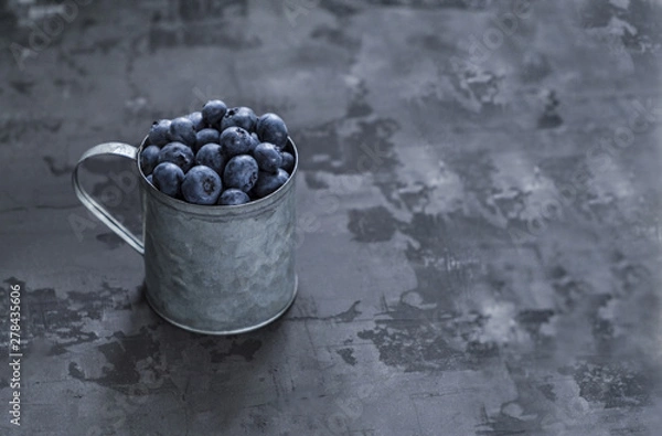 Obraz Fresh blueberry berries in an iron mug on a dark background. Copy space