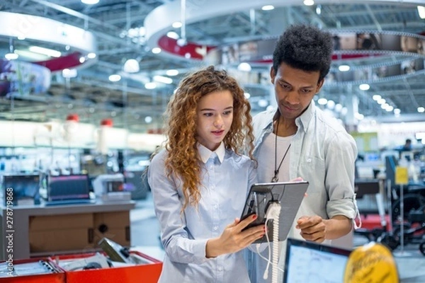 Fototapeta Red-haired girl with glasses with an African guy in an electronics store standing in front of the stand choosing a tablet computer to buy