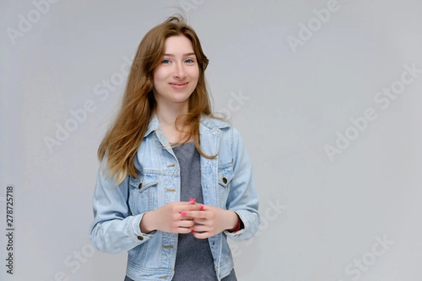 Fototapeta Portrait to waist of a young pretty brunette girl woman with beautiful long hair on a white background in a jacket from jeans. He talks, smiles, shows his hands with emotions in various poses.