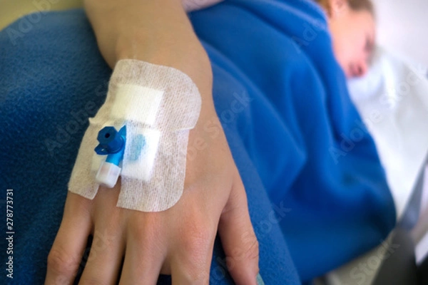 Obraz Patient's hand with blue Infusion catheter on blue blanket background. Sick woman with catheter lies on the couch. Catheter closed up