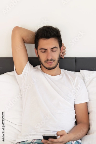 Fototapeta Young handsome man sitting on the bed. He uses a mobile phone. The problem of dependence on mobile social networks.