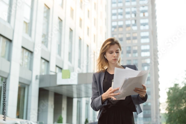 Obraz Concentrated stylish businesswoman standing on city street with documents reading them. Successful female manager dressed in elegant suit checking accounting on paper during coffee break outdoors.