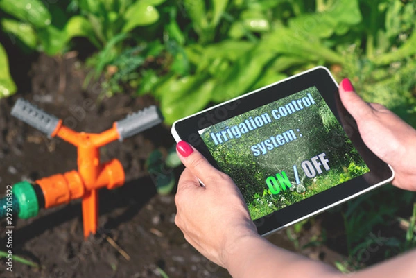 Fototapeta Woman is holding in hand a tablet computer with a program of irrigation control system on a garden sprinkler background.