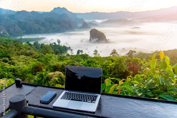Fototapeta A laptop, mobile  on wooden table with sunrise and mountain fog background in morning. A start of new day. Freelance business concept.