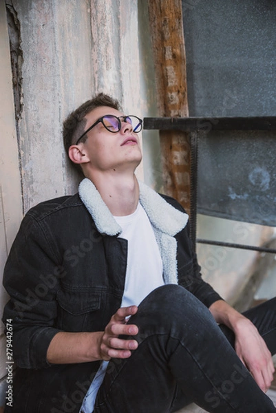 Fototapeta a young rebel in a jacket and jeans on the background of an old crumbling building with glasses and brutal hair
