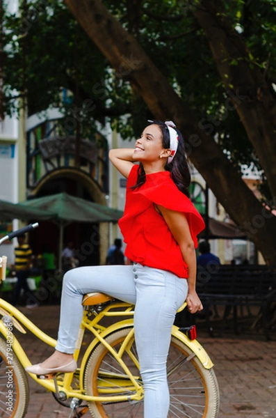Obraz Outdoor portrait of young beautiful girl 19 to 25 years old posing in street. Brunette. riding a retro styled bicycle. Wearing red blouse With a penetrating camera look. 