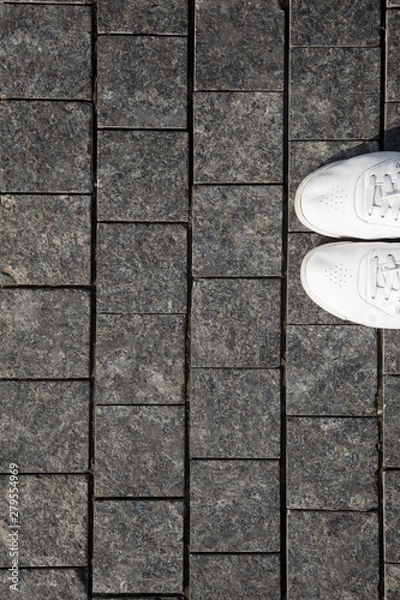 Fototapeta Top view of white sneakers and gray cobblestone pavement. Gray stone pavement background.