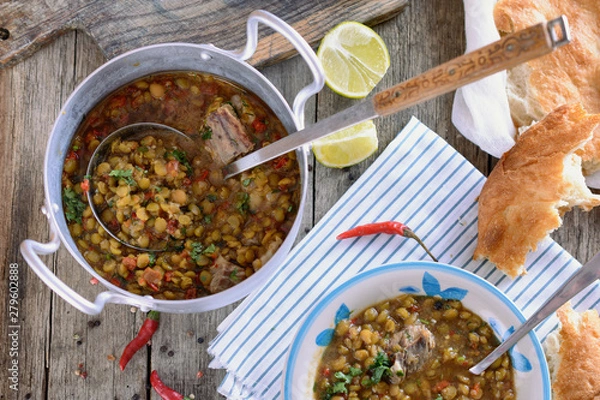 Fototapeta Mixed legumes and peas soup with meat in a vintage aluminum pan with a ladle for serving on an old wooden table next to a bowl with this soup, slices of tortilla, lime and chili pepper.