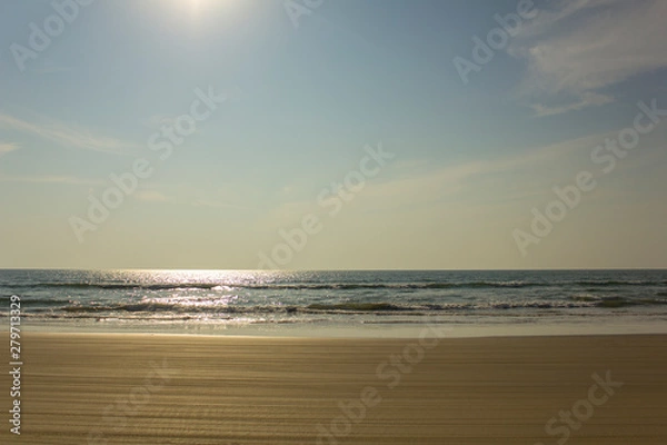Fototapeta yellow sandy beach with motorcycle tire tracks against the backdrop of sea waves under a clear blue sky with the sun